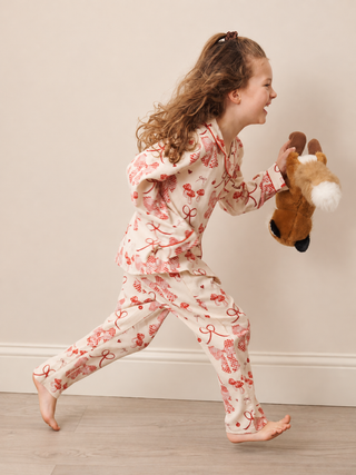 A young girl with long curly brown hair is captured mid-run wearing a cream-coloured pyjama set with a red bow and ribbon pattern. She is holding a plush toy in a playful indoor setting with a light beige wall and wood floor.