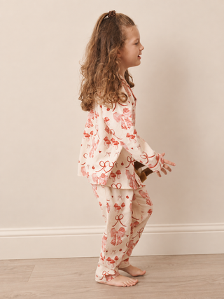 A young girl with long wavy brown hair stands in profile wearing a cream-colored pyjama set with a red bow and ribbon pattern. She is standing barefoot on a light wood floor against a plain beige wall.