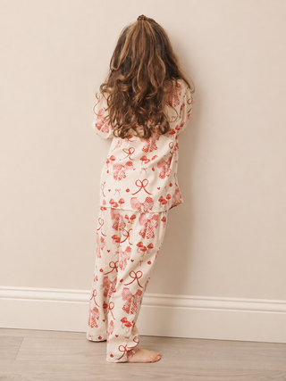 A child standing with their back to the camera, wearing a two-piece pyjama set with a red bow and strawberry pattern on a cream background, set against a plain beige wall and light wood floor.