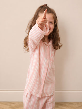 A young girl with curly brown hair pointing her finger while wearing a pink and white vertically striped pajama set against a neutral beige wall.