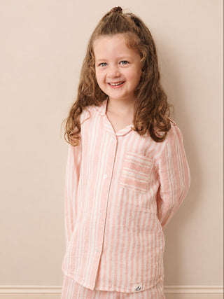 A young girl with long light brown hair stands against a beige wall wearing a pink and white vertically striped pajama set. The set includes a long-sleeve button-up top with a chest pocket and matching pants.