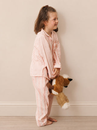 A young girl stands in profile wearing pink and white vertically striped pyjamas and holding a brown plush fox toy against a plain beige wall.