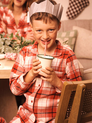 A young boy wearing red and white plaid pyjamas and a paper crown sips from a white mug with a red-and-white striped straw in a cozy indoor setting. A woman in matching plaid pajamas is blurred in the background.