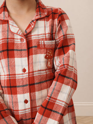 A close-up of a person wearing a red, white, and black plaid pajama shirt with a mushroom patch on the pocket, set against a neutral beige background.