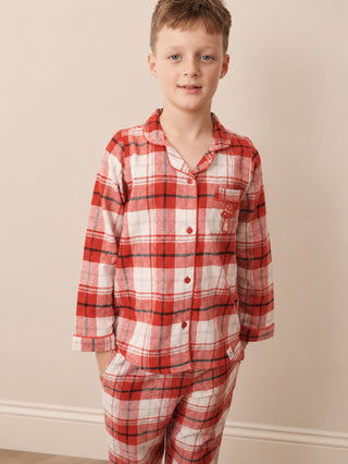 A young boy wearing a red, white, and black plaid tartan pyjama set, standing against a plain beige wall.