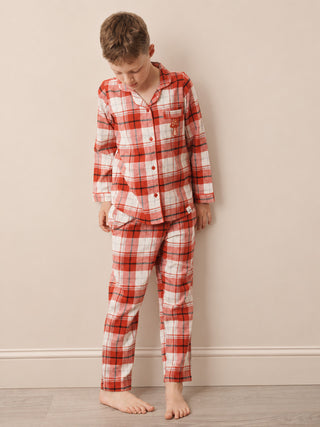 A young boy wearing a red, white, and black plaid pajama set, standing barefoot against a plain beige wall.