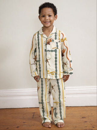 A young child wearing a two-piece pyjama set with a vertical striped pattern in olive green, mustard yellow, and white, decorated with illustrations of sea life like starfish and crabs, standing against a plain off-white wall.
