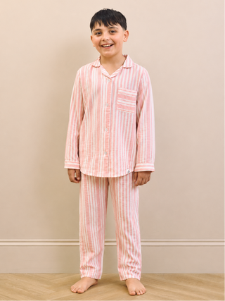 A young boy stands barefoot against a beige wall wearing a pink and white vertically striped pajama set with a long-sleeve button-up shirt and matching pants.