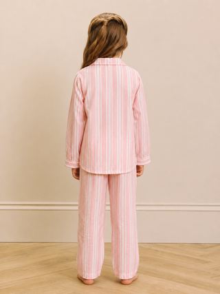 A child standing with their back to the camera, wearing a light pink and white vertically striped pajama set consisting of a long-sleeve top and matching pants, in a room with a beige wall and light wood floor.