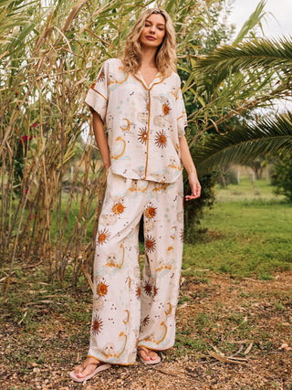 A woman standing outdoors wearing a short-sleeved, cream-coloured pyjama set with a celestial print of suns, moons, and clouds.
