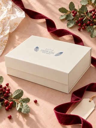 A cream-colored gift box with a blue printed logo reading 'Kings Christmas Bed & Break' sits on a dusty pink background, styled with a dark red velvet ribbon, green eucalyptus leaves, red berries, and a blank gift tag.
