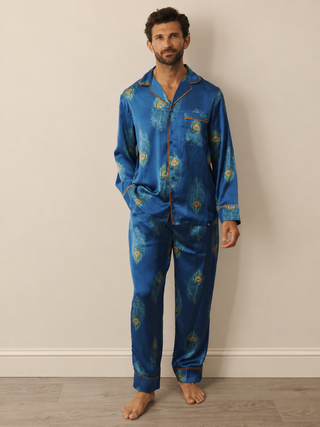 A man wearing a navy blue satin pyjama set with a gold and green peacock pattern, standing against a plain beige wall on a light wood floor.