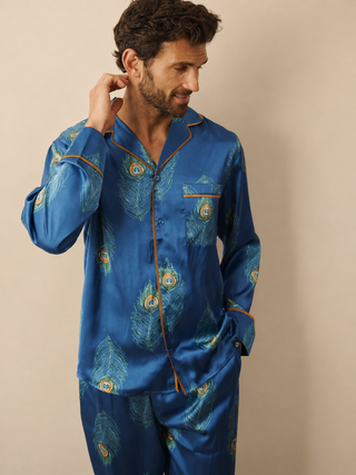A man wearing a navy blue satin pyjama set with a peacock feather pattern and gold trim, posing against a solid beige background.