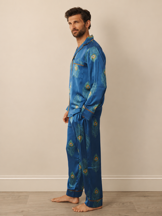A man wearing a navy blue pyjama set with a gold and green paisley pattern, standing in profile against a beige wall and light wood floor.