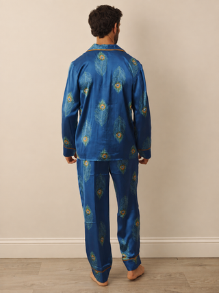 A man viewed from behind wearing a blue satin pyjama set with a gold and green peacock feather pattern and gold piping trim, standing against a beige wall.