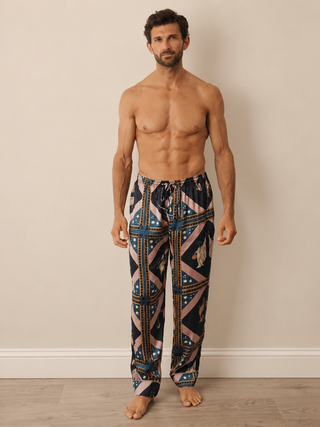 A shirtless model stands barefoot against a plain beige wall, wearing loose patterned pyjama bottoms with a geometric print in navy, pink, blue, and beige.