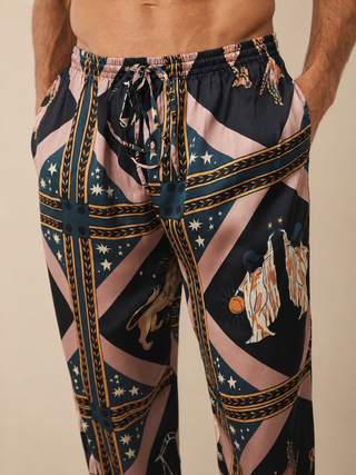 A close-up of a person wearing patterned drawstring pants. The pants have a complex graphic print featuring diagonal pink stripes, dark navy sections with white stars, gold decorative borders, and illustrations of figures in white robes.