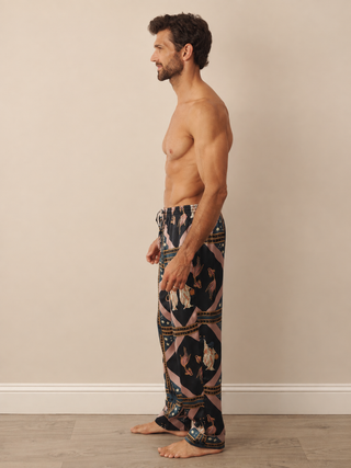 A man stands in profile wearing loose patterned pyjama pants with a geometric and floral design in black, pink, beige, and blue, against a plain beige wall and light wood floor.