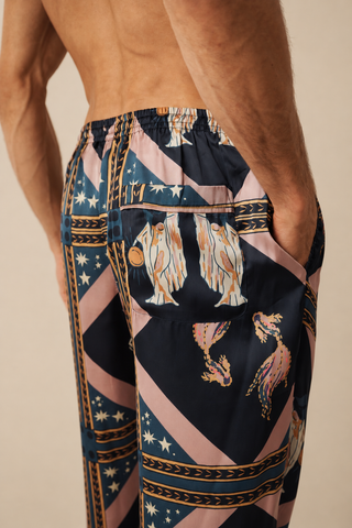 A close-up of a man wearing patterned pyjama trousers, showing the back and side of the garment. The pants have a dark navy base with pink diagonal stripes, gold decorative borders, and illustrations of winged figures and birds.