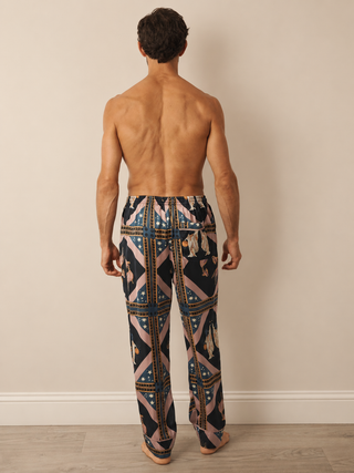 A man viewed from behind wearing patterned pyjama lounge pants with a geometric design in navy, pink, beige, and gold, standing against a plain beige wall.