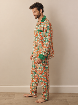 A man wearing a two-piece pyjama set with a colourful geometric pattern in orange, green, and white, featuring solid green collar and cuff accents, standing against a beige wall.