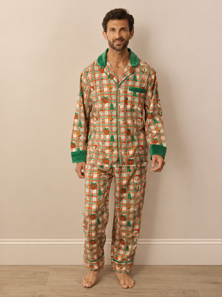 A man wearing a long-sleeve Christmas pyjama set with a brown, orange, and green geometric pattern and solid green collar and cuffs, standing against a beige wall.