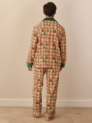 A person standing with their back to the camera wearing a matching two-piece pyjama set with a festive holiday print of Christmas trees, ornaments, and stockings on an orange and white plaid background, with green trim.