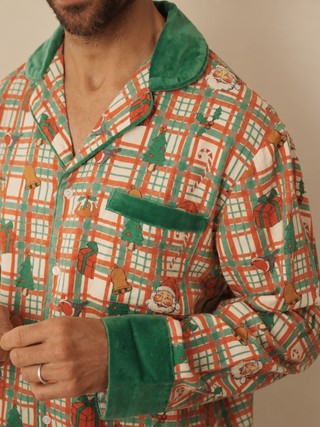 A man wearing a long-sleeve pyjama shirt with a festive Christmas plaid pattern in green, orange, and white, featuring green velvet trim on the collar, pocket, and cuffs.