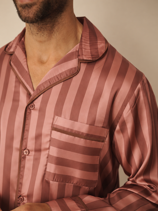 A close-up of a man wearing a satin pyjama shirt with vertical dusty rose and brown stripes, featuring a collar, button-down front, and a chest pocket.