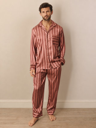 A man wearing a two-piece satin pyjama set with vertical stripes in dusty mauve and rose , standing against a beige wall and light wood floor.