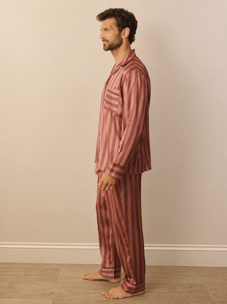 A man stands in profile wearing a matching two-piece pyjama set with vertical stripes in dusty rose and dark red, set against a plain beige wall and light wood floor.
