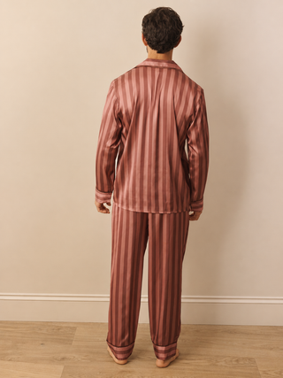 A man viewed from behind wearing a matching two-piece pyjama set with vertical stripes in dusty rose and dark brown, standing on a light wood floor against a beige wall.