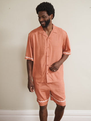 Model wearing a rust-coloured men’s linen-blend short pyjama set with a button-through short-sleeve shirt and matching shorts, finished with pale pink ric-rac trim, photographed barefoot against a neutral wall.
