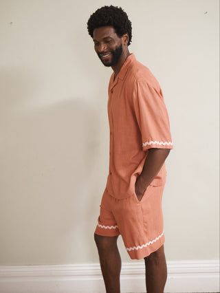 Model wearing a rust-coloured men’s linen-blend short pyjama set with a button-through short-sleeve shirt and matching shorts, finished with pale pink ric-rac trim, photographed barefoot against a neutral wall.