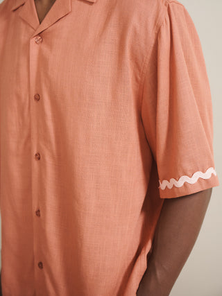 Model wearing a rust-coloured men’s linen-blend short pyjama set with a button-through short-sleeve shirt and matching shorts, finished with pale pink ric-rac trim, photographed barefoot against a neutral wall. Detail shot
