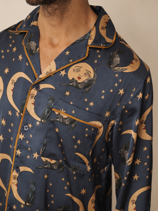 A close-up of a man wearing a navy blue pyjama shirt with a whimsical print of black cats on crescent moons and stars, featuring gold trim and a gold pocket.