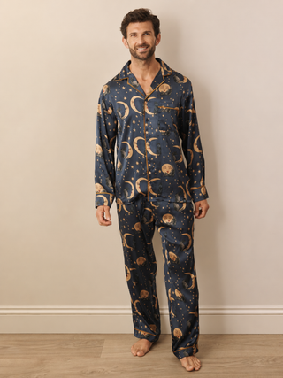 A man wearing a navy blue pyjama set with a gold celestial pattern of crescent moons, stars, and planets, standing against a beige wall and light wood floor.
