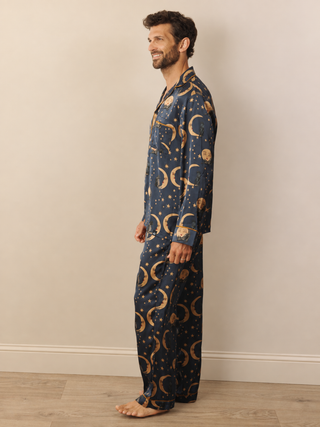 A man wearing a navy blue pyjama set with a gold celestial print of crescent moons and stars, standing in profile against a beige wall and light wood floor.