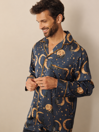 A man wearing a navy blue pyjama set with a gold celestial print of crescent moons, stars, and sleeping faces, shown against a plain beige background.