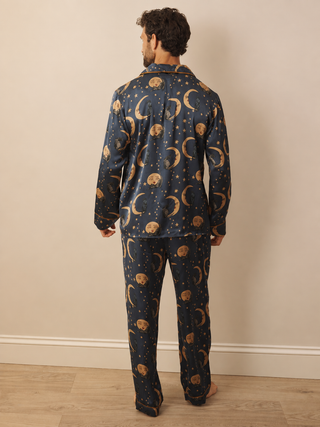 A man viewed from behind wearing a navy blue pyjama set with a gold celestial print of crescent moons, stars, and planets, standing against a beige wall.