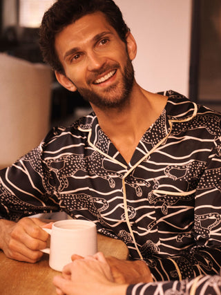 A smiling man wearing a black and white patterned pyjama shirt sits at a table holding a white mug.