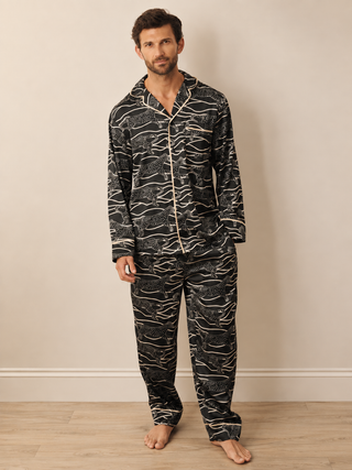 A man modelling a two-piece pyjama set with a black and beige wavy abstract cheetah pattern, standing against a neutral beige wall and light wood floor.