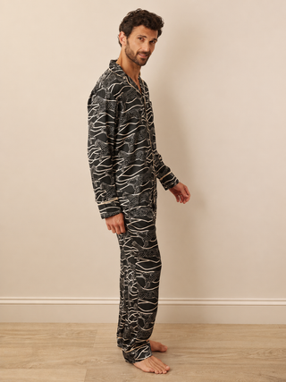 A man wearing a black and white abstract patterned long-sleeve pyjama set, standing barefoot against a beige wall and light wood floor.