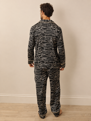 A man viewed from behind wearing a matching long-sleeve pyjama set with an abstract black and white pattern, standing against a plain beige wall on a light wood floor.