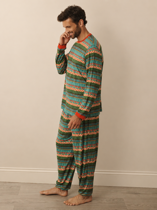 A man wearing a two-piece long-sleeve pyjama set with a green, teal, and orange geometric pattern, standing barefoot against a beige wall.