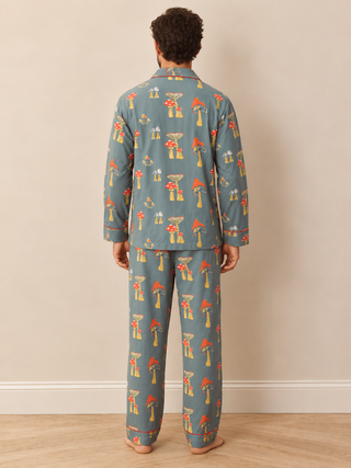 A man viewed from behind wearing a matching long-sleeve pyjama set with an all-over mushroom print in muted blue-gray, red, orange, and yellow, standing against a plain beige wall on a light wood floor.