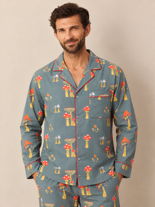 A man wearing a matching long-sleeve pyjama set with a blue-grey base and an all-over red, yellow, and white mushroom print, standing against a plain beige studio background.