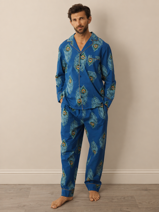 A man wearing a two-piece pyjama set with a vibrant blue base and a detailed peacock feather print, standing against a neutral beige wall and light wood floor.