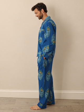 A man wearing a blue pyjama set with a peacock feather print, standing in profile against a beige wall and light wood floor.