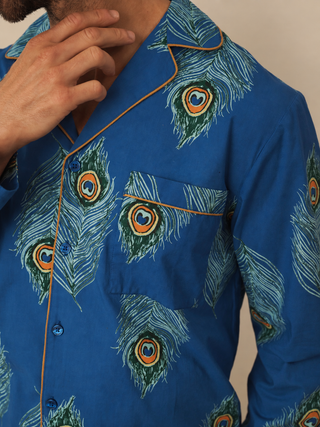 A close-up of a men's long-sleeve pyjama shirt featuring a vibrant blue base with a green and white peacock feather print and thin orange piping along the collar, placket, and pocket.